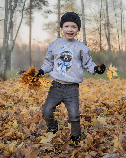 A smiling young boy modeling the grey Annette Thomé-Marttinen x Novapuro Winter Raccoon youth oversize sweatshirt, enjoying the outdoors in autumn.