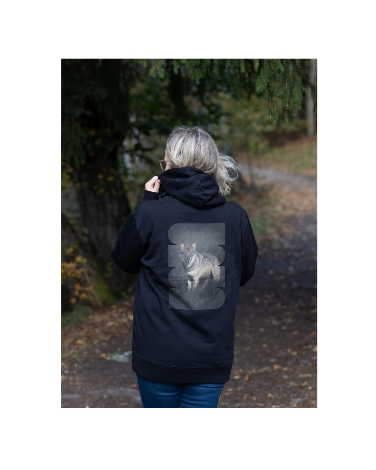 Female model in a forest wearing a black unisex organic cotton hoodie with a striking Dark Alpha Wolf back print. Sustainable Nordic streetwear by Auli Leivonen x Novapuro.
