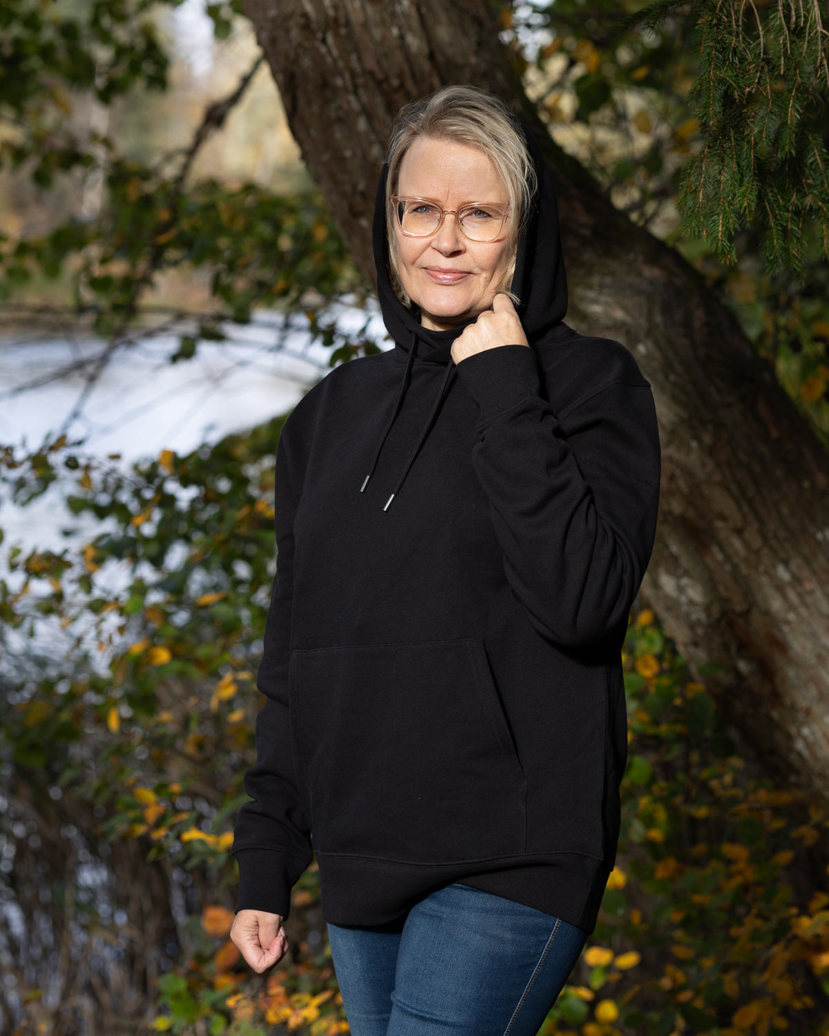 Auli Leivonen wearing a black Novapuro organic cotton hoodie with a majestic Nordic bear photography print on the back, standing outdoors by a lake.