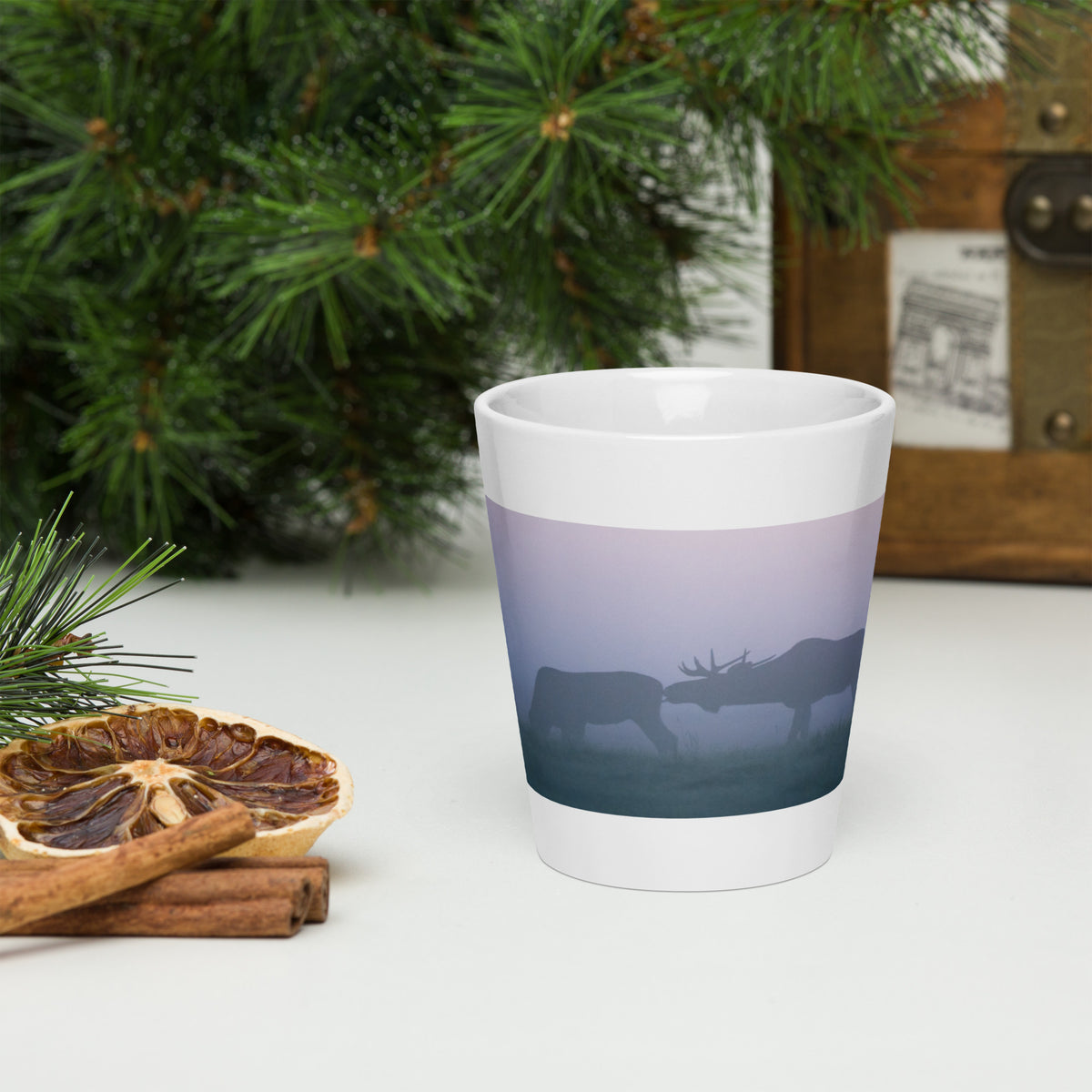 A white ceramic cone-shaped latte mug featuring an original photograph of two moose in the mist by Mikko Monto, set in a cozy rustic environment