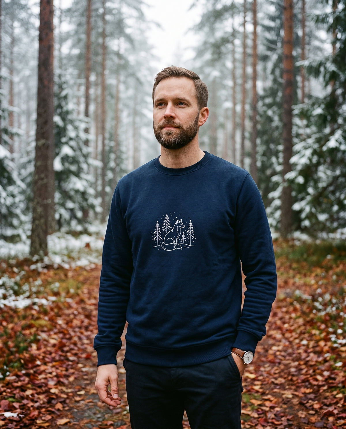 A bearded male model wearing a sustainable Novapuro navy blue organic sweatshirt with a minimalist white arctic fox forest design, standing in a snowy Nordic woods.