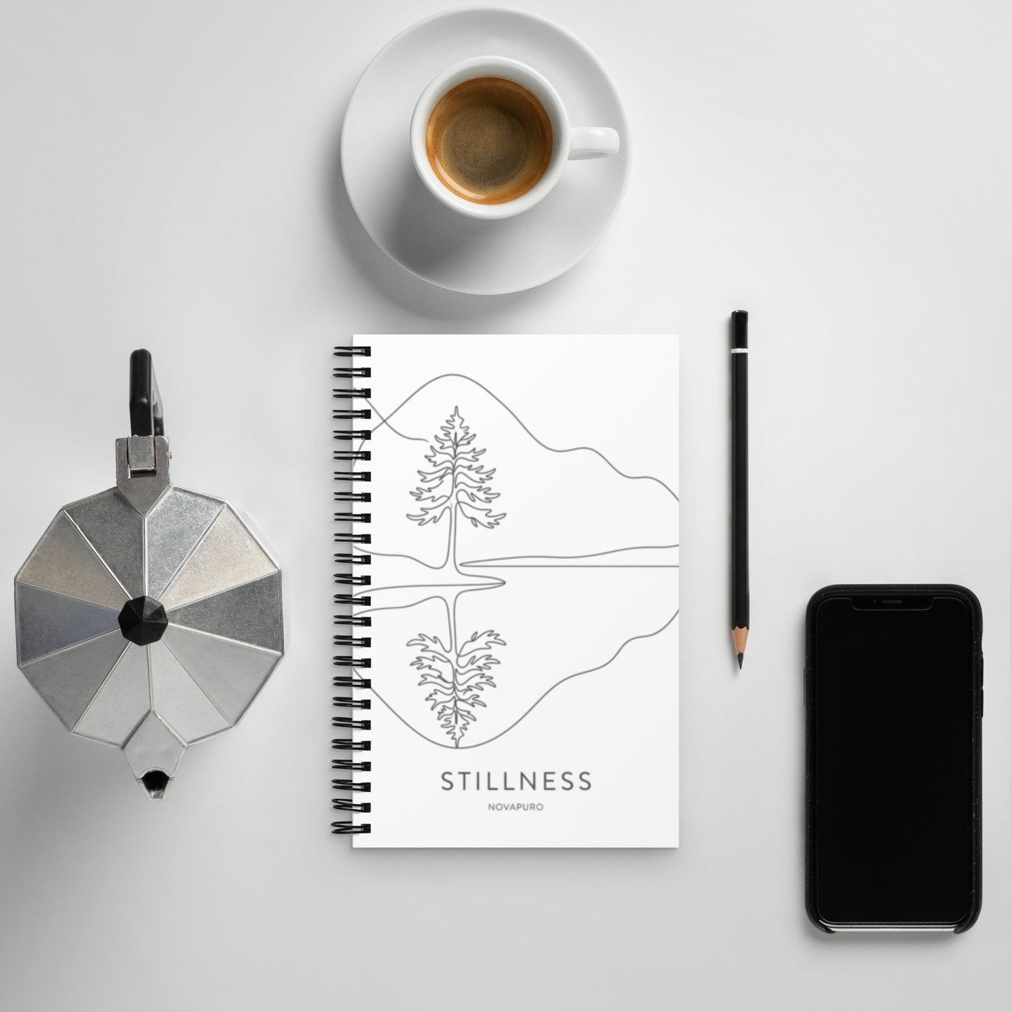 A white spiral-bound notebook laying flat on a desk next to a coffee cup, featuring a minimalist charcoal grey line art illustration of a Nordic lake reflecting a pine tree with the text "STILLNESS".