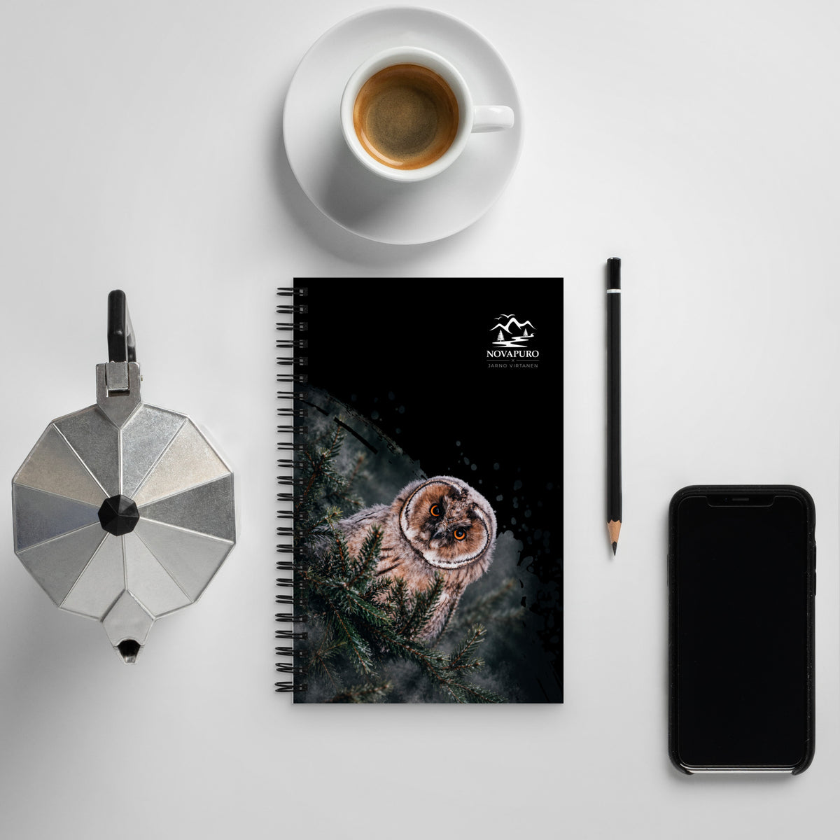 A black wire-bound spiral notebook featuring a Nordic long-eared owl chick print by Jarno Virtanen, placed on a white desk next to a coffee cup.