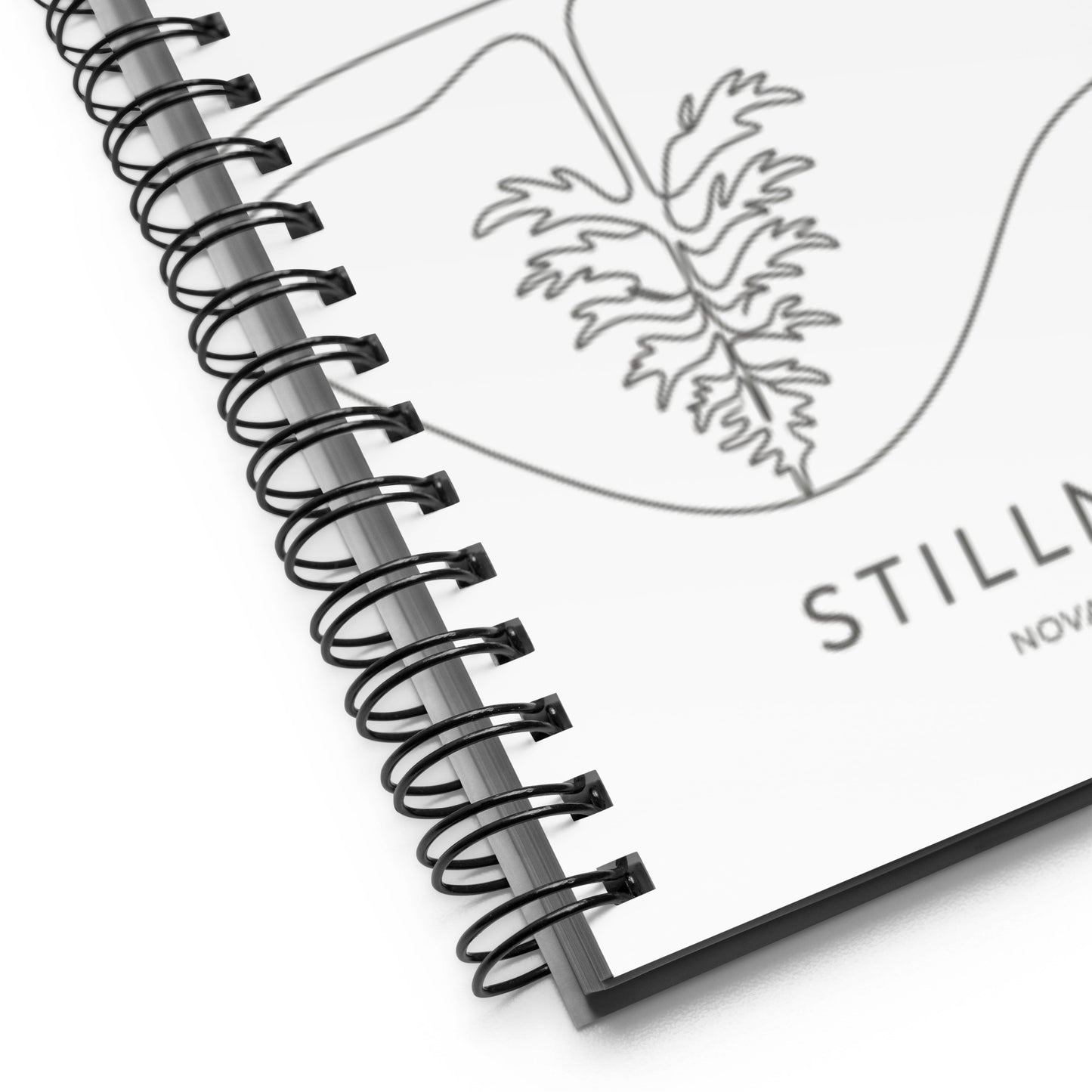 A white spiral-bound notebook laying flat on a desk next to a coffee cup, featuring a minimalist charcoal grey line art illustration of a Nordic lake reflecting a pine tree with the text "STILLNESS".