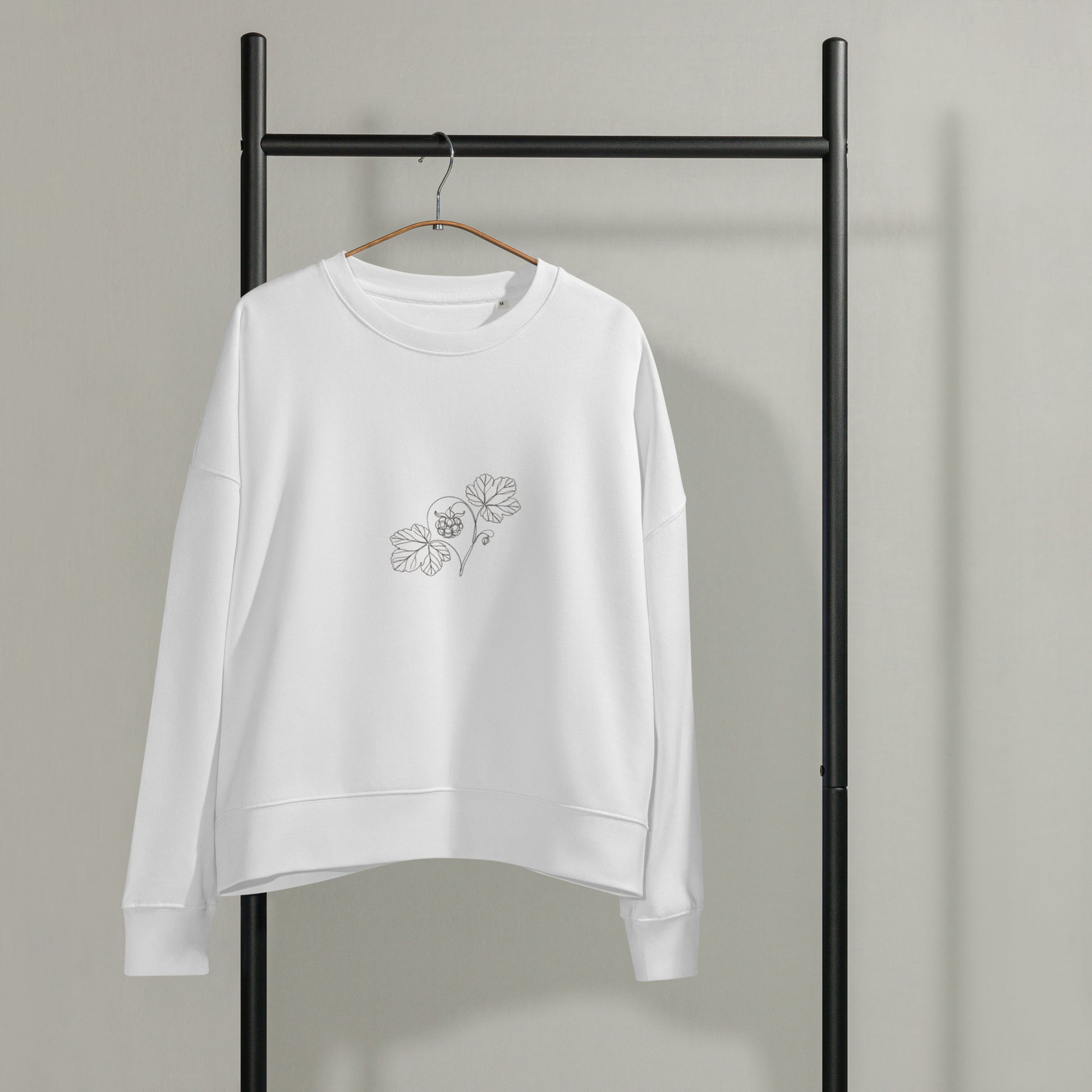 White Novapuro women's organic cotton crewneck sweatshirt hanging on a black garment rack, featuring a minimalist botanical cloudberry line art illustration on the left chest